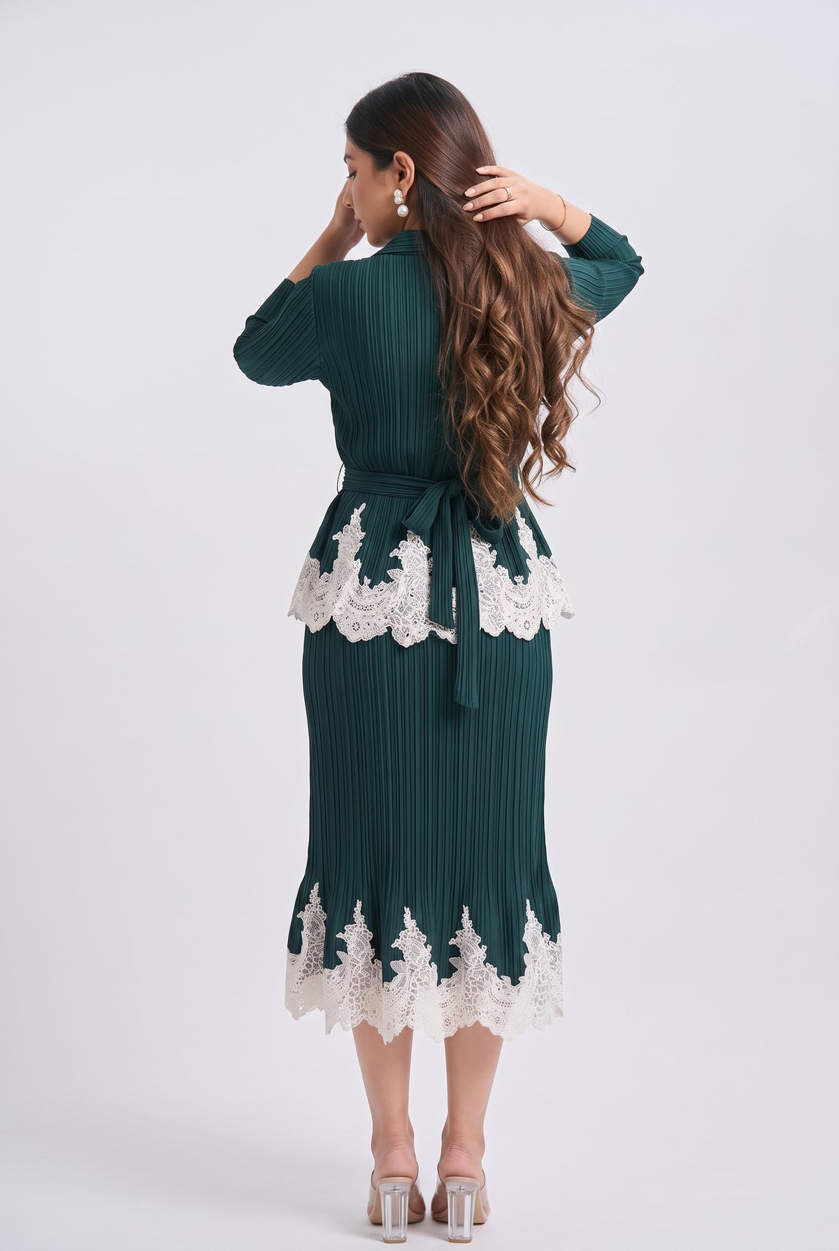 A woman shown from the back wearing a dark green pleated midi dress with white lace trim at the waist and hem, paired with clear high-heeled sandals, against a light gray studio background.