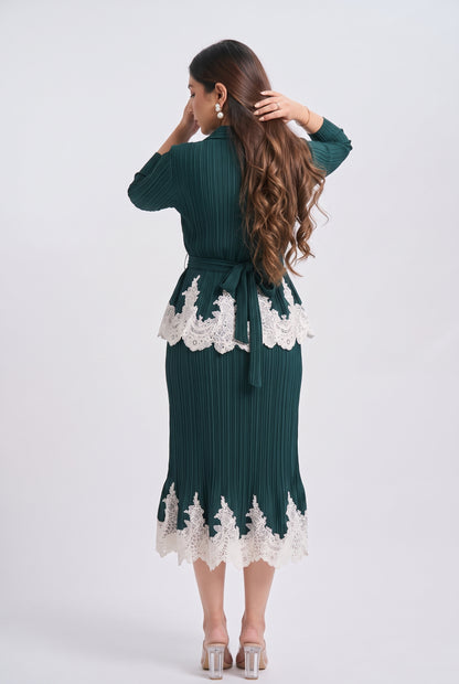 A woman shown from the back wearing a dark green pleated midi dress with white lace trim at the waist and hem, paired with clear high-heeled sandals, against a light gray studio background.