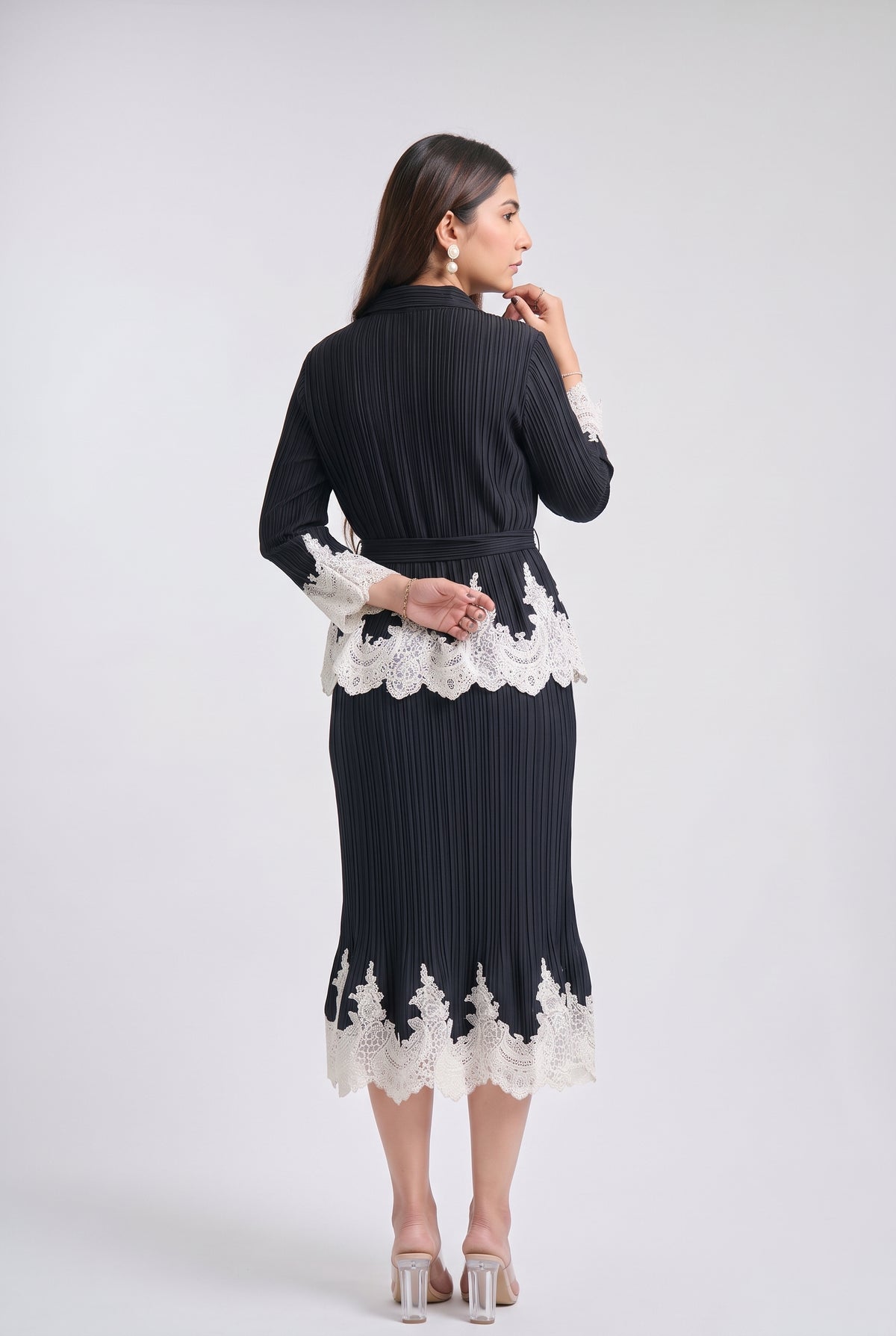 A woman is shown from the back wearing a black pleated long-sleeve midi dress with white lace trim at the waist and hem, paired with clear high-heeled sandals, against a light gray studio background.