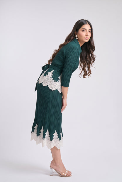 A woman modeling a dark green pleated midi dress with white lace trim details, shown from a side-back angle against a light gray studio background.