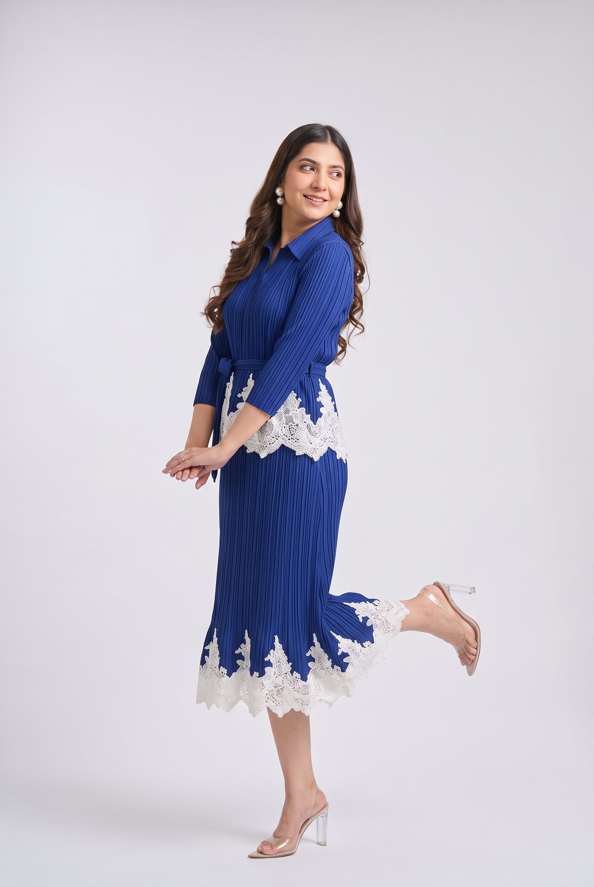A woman modeling a royal blue pleated midi dress with white lace trim at the waist and hem, paired with clear high-heeled sandals, against a light gray studio background.