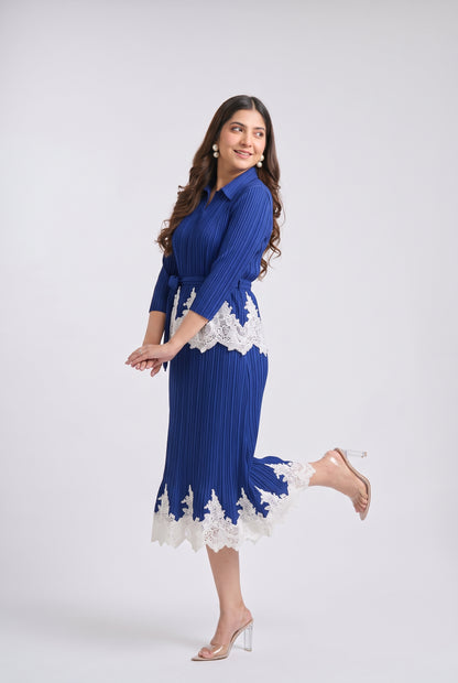 A woman modeling a royal blue pleated midi dress with white lace trim at the waist and hem, paired with clear high-heeled sandals, against a light gray studio background.