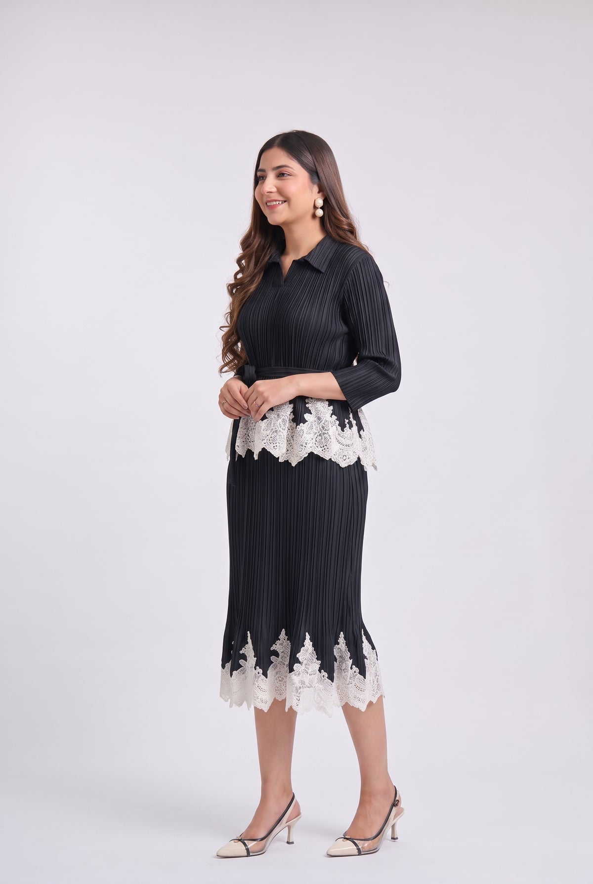 A woman modeling a black pleated midi dress with white lace trim at the waist and hem, paired with beige high-heeled sandals, against a light gray studio background.