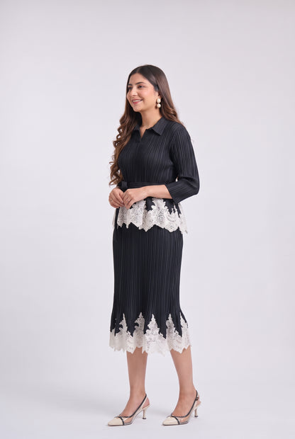 A woman modeling a black pleated midi dress with white lace trim at the waist and hem, paired with beige high-heeled sandals, against a light gray studio background.