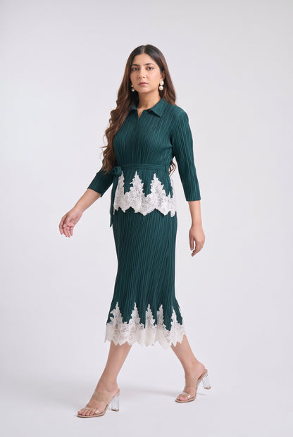 A woman models a dark green pleated midi dress with white lace trim at the waist and hem, paired with clear high-heeled sandals, against a light gray studio background.