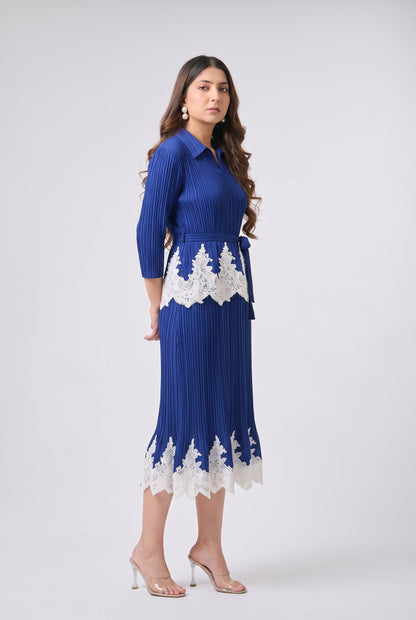 A woman modeling a royal blue pleated midi dress with white lace trim at the waist and hem, paired with clear high-heeled sandals, against a light gray studio background.