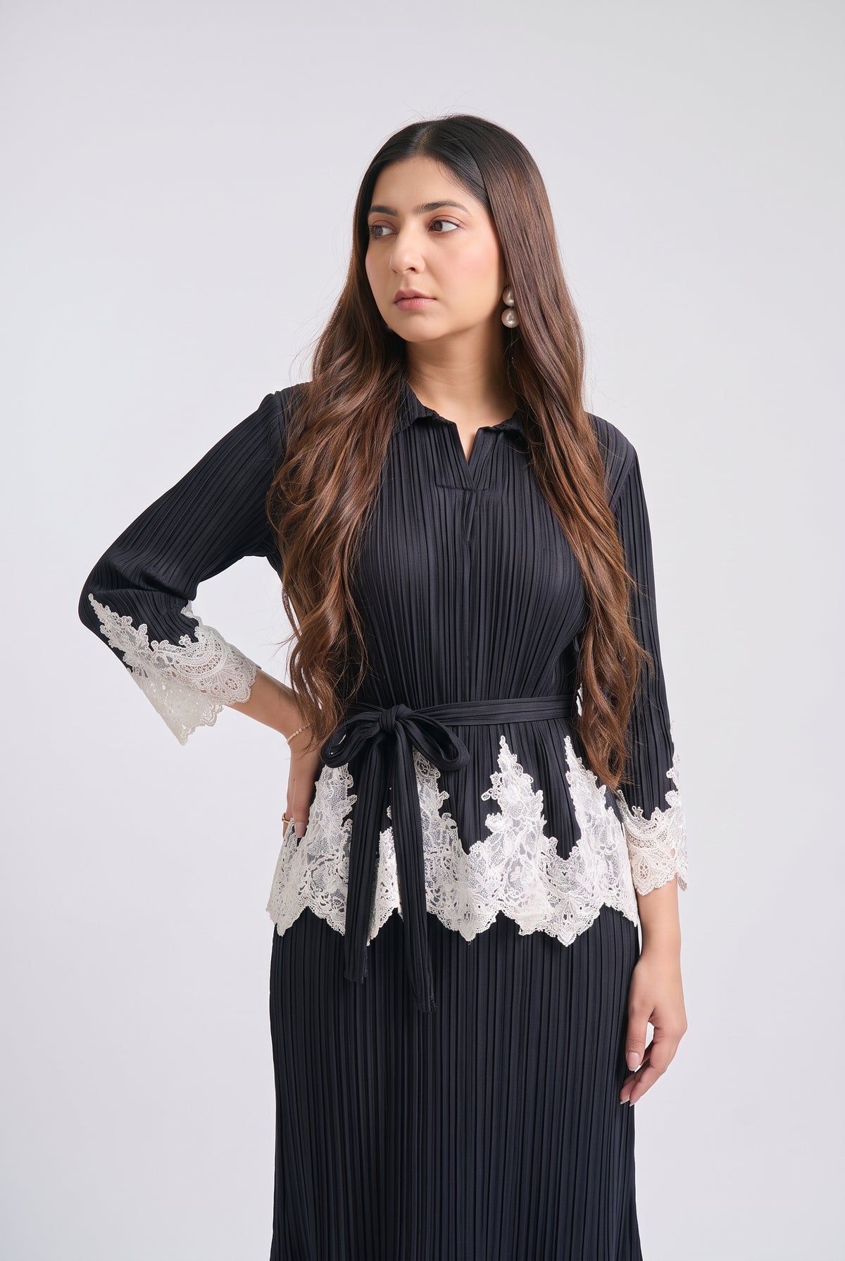 A woman modeling a black pleated long-sleeve dress with white lace trim at the cuffs and waist, tied with a matching black belt, against a light gray studio background.