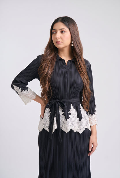 A woman modeling a black pleated long-sleeve dress with white lace trim at the cuffs and waist, tied with a matching black belt, against a light gray studio background.