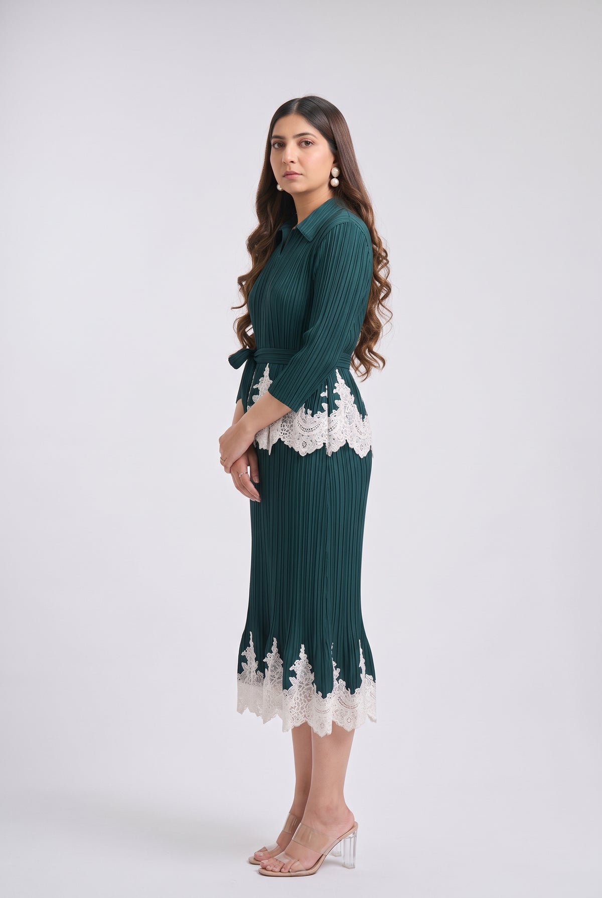 A woman modeling a dark teal green pleated midi dress with white lace trim at the waist and hem, paired with clear high-heeled sandals, against a light gray studio background.