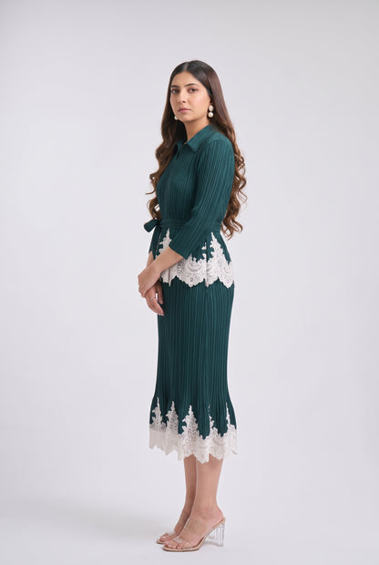 A woman modeling a dark teal green pleated midi dress with white lace trim at the waist and hem, paired with clear high-heeled sandals, against a light gray studio background.