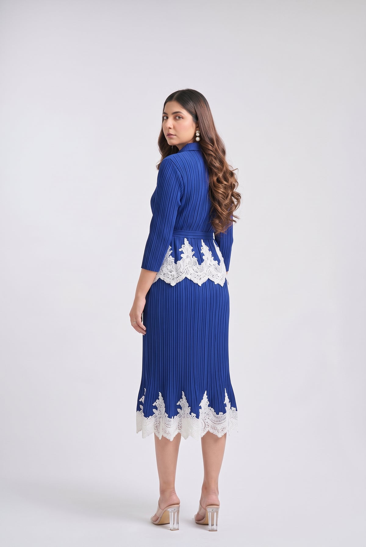A woman modeling a royal blue pleated midi dress with white lace trim at the waist and hem, shown from the back against a light gray studio background.