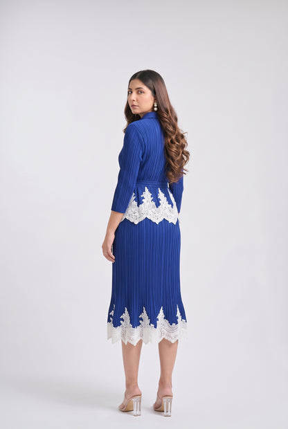 A woman modeling a royal blue pleated midi dress with white lace trim at the waist and hem, shown from the back against a light gray studio background.