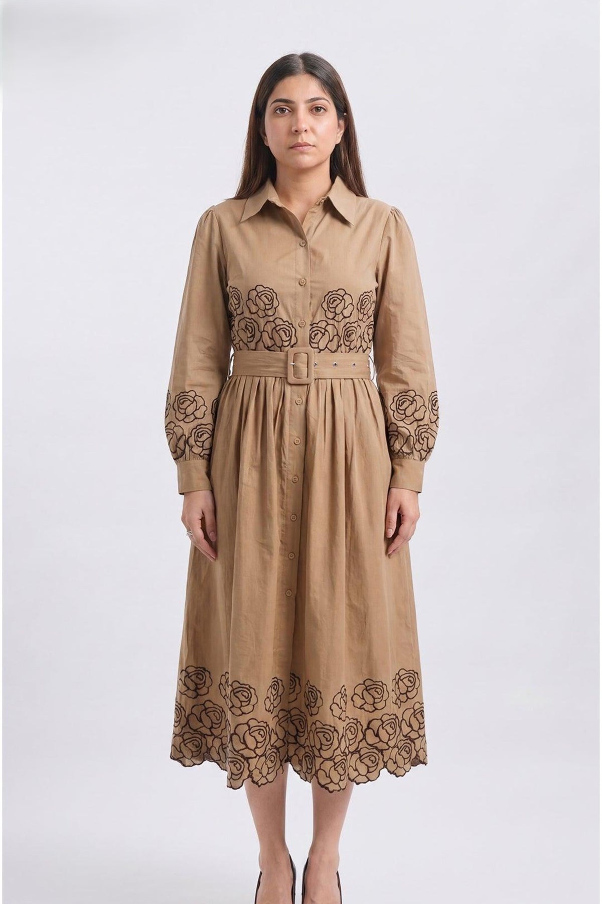A woman modeling a tan long-sleeve midi shirt dress with dark brown floral lace embroidery at the chest, cuffs, and hem, set against a plain light gray studio background.