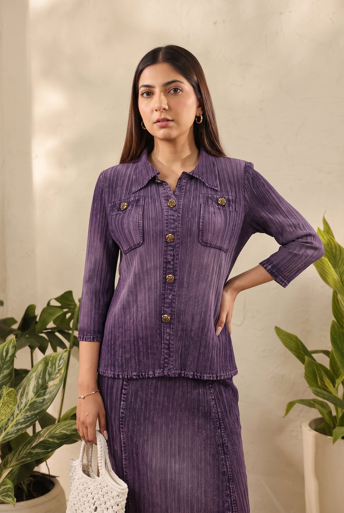 A woman modeling a matching purple textured two-piece set consisting of a long-sleeve button-front shirt jacket and a skirt, styled with a white woven handbag in an indoor setting with potted plants.