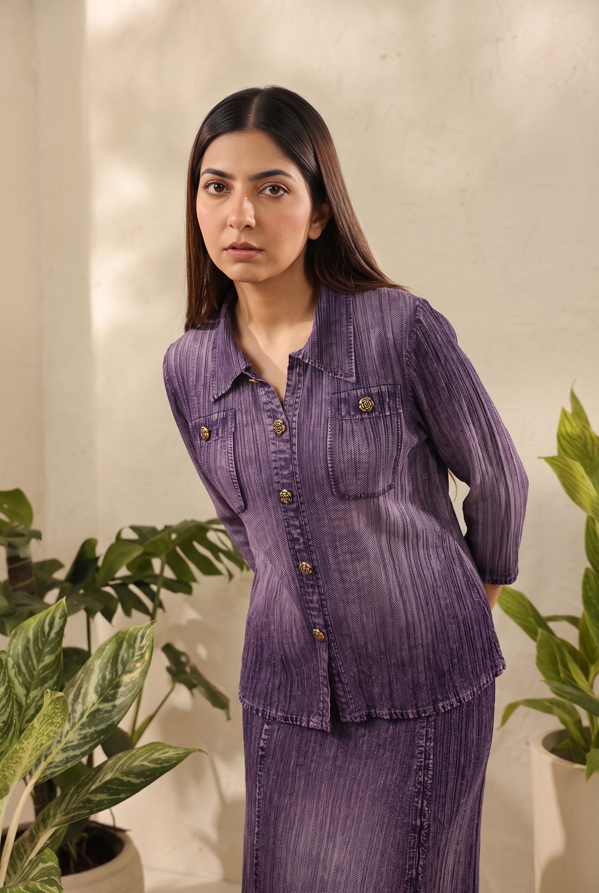 A woman wearing a matching purple textured two-piece set consisting of a long-sleeve button-front top and a skirt, standing in front of a neutral wall with potted plants.