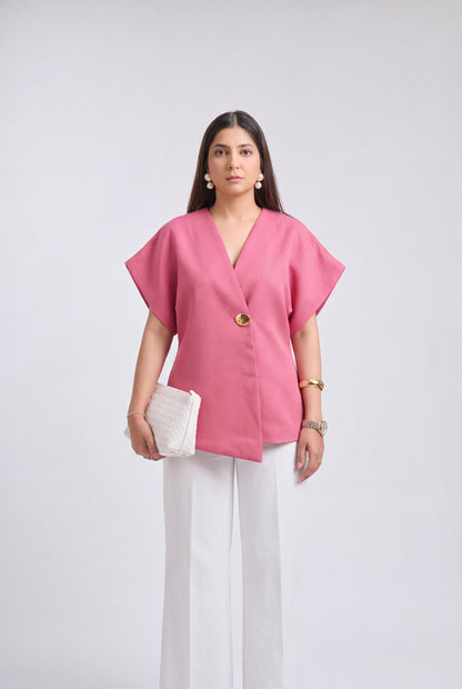 A woman modeling a pink short-sleeve wrap-style top with a single gold button, paired with white wide-leg pants and holding a white textured clutch, against a light gray studio background.