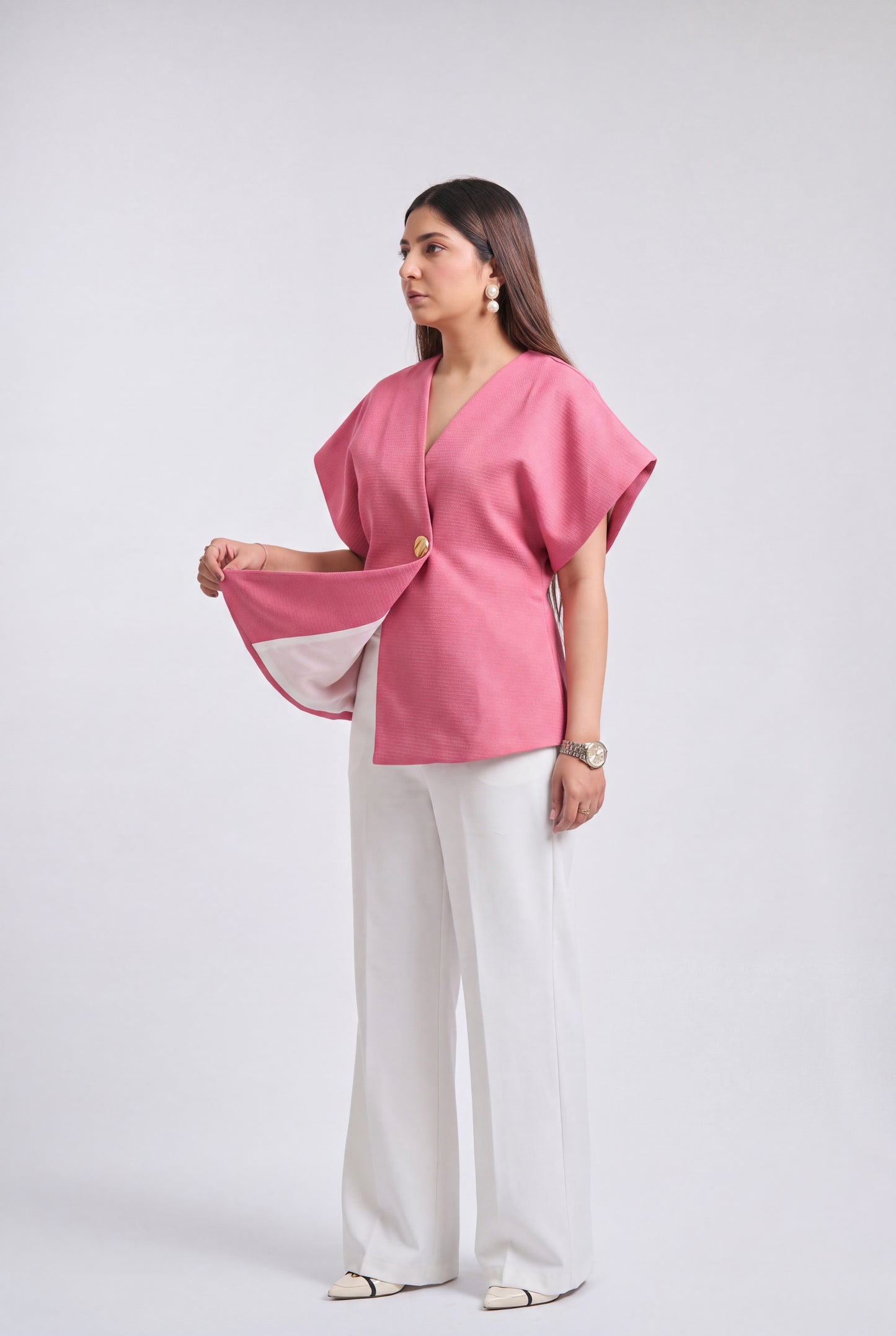 A woman modeling a pink short-sleeve wrap-style top with white wide-leg trousers, standing against a light gray studio background.