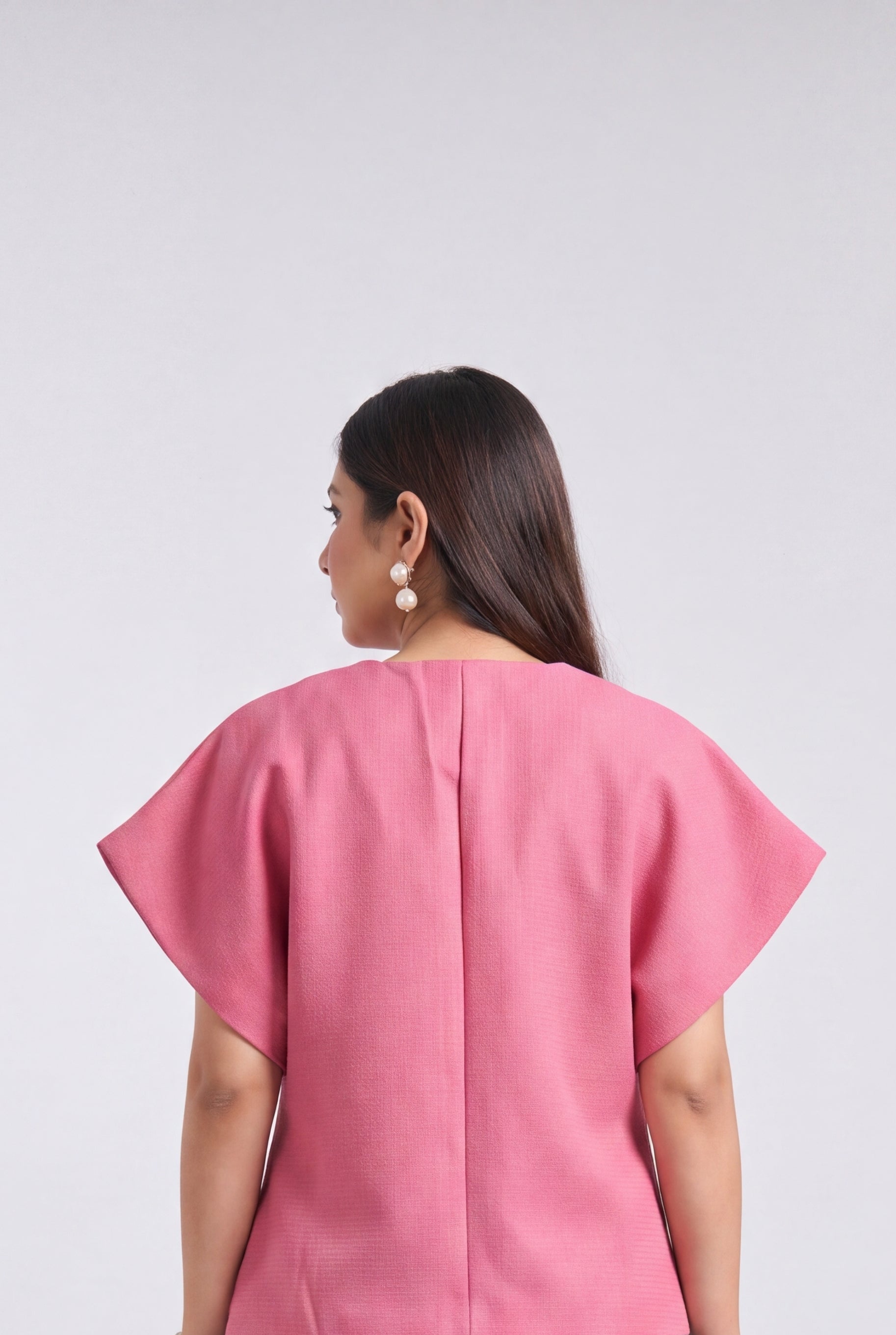 A woman viewed from the back wearing a pink short-sleeve top with a structured, slightly draped back and blue jeans with white heart embroidery on the back pockets, set against a plain light gray studio background.