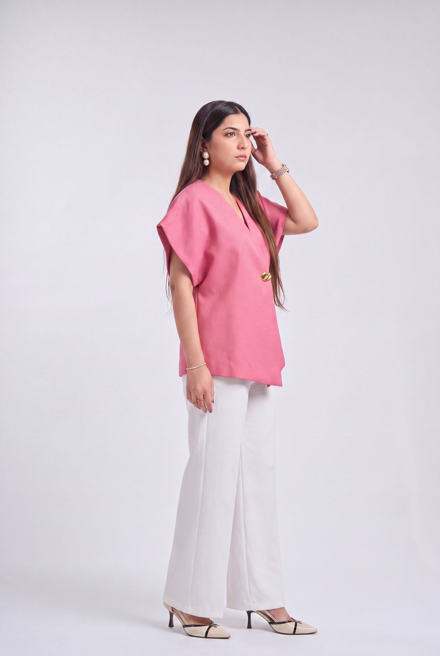 A woman modeling a pink short-sleeve top with a V-neck and gold button detail, paired with white wide-leg pants and beige heeled sandals, against a light gray studio background.