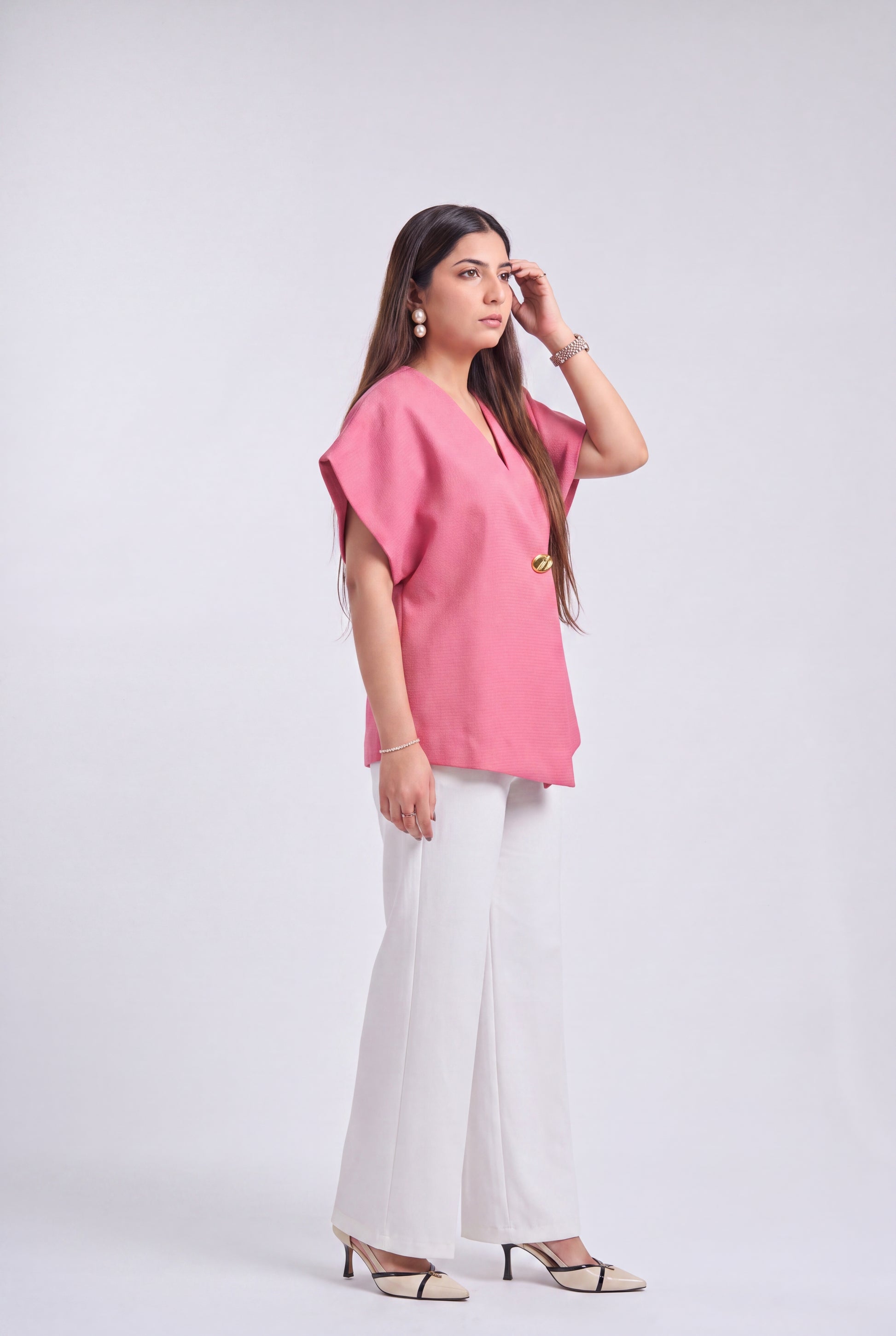 A woman modeling a pink short-sleeve top with a V-neck and gold button detail, paired with white wide-leg pants and beige heeled sandals, against a light gray studio background.