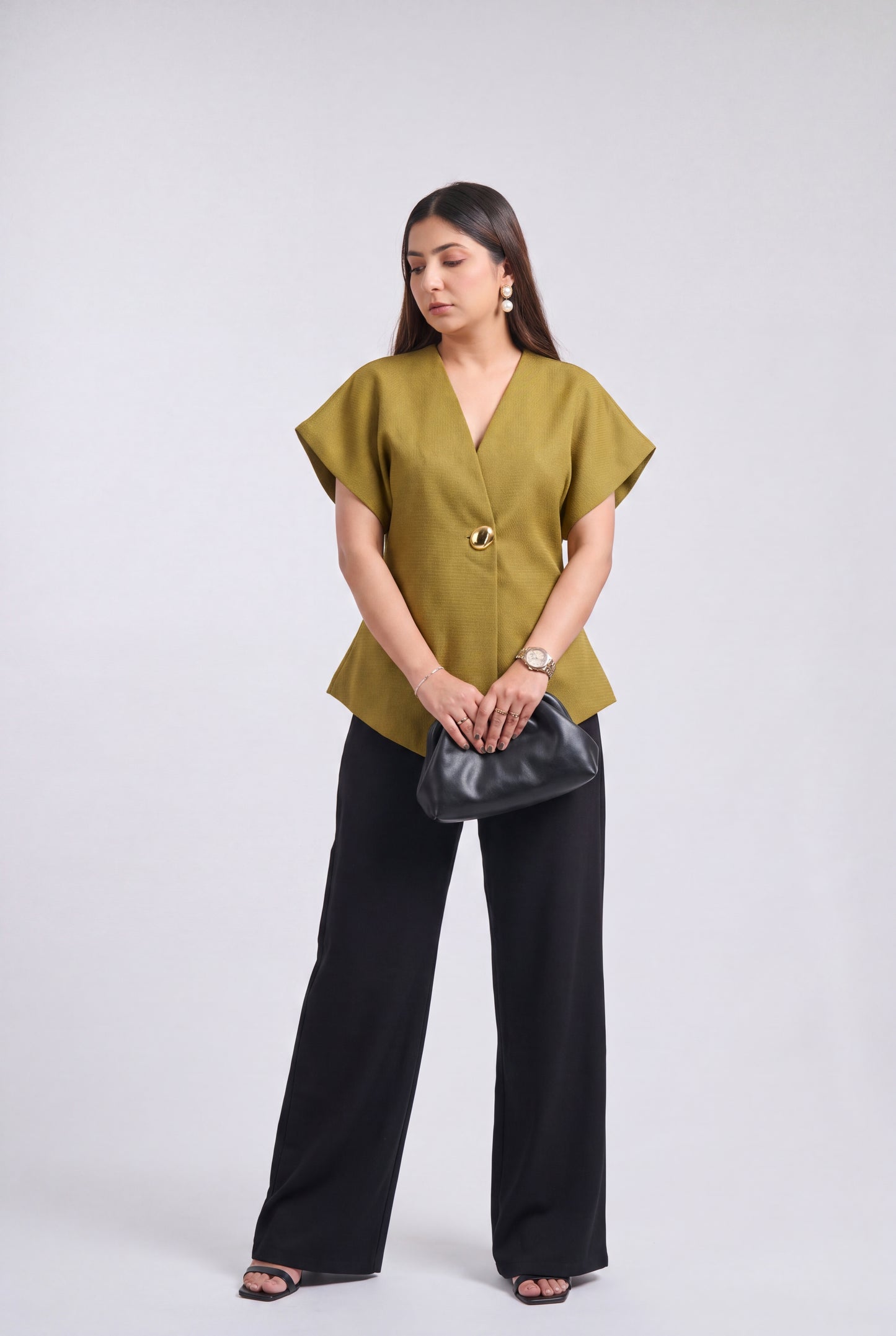A woman modeling a short-sleeve olive green wrap-style blouse with a single decorative button, paired with wide-leg black pants and holding a black clutch bag, against a light gray studio background.