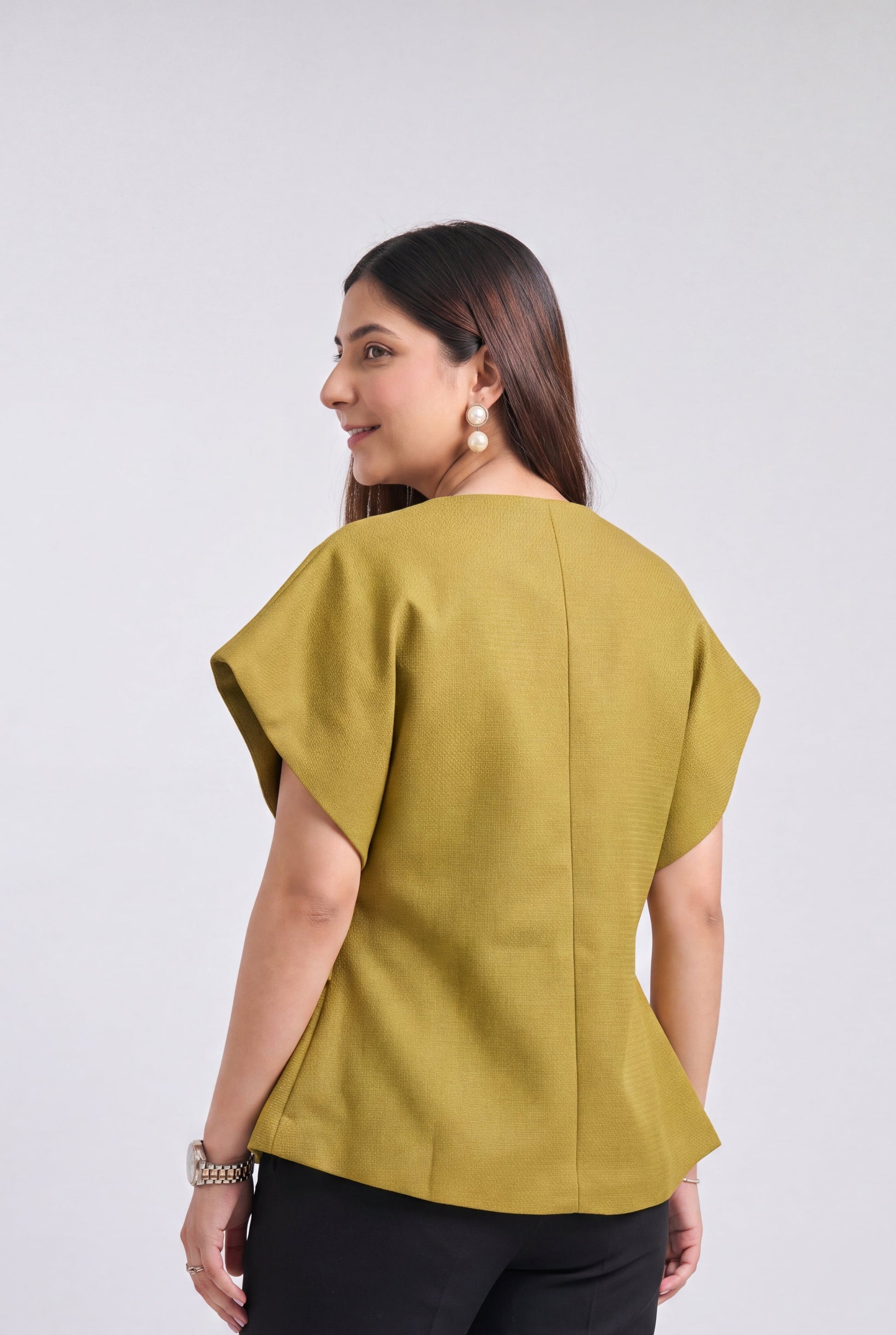 A woman shown from the back wearing a mustard yellow short-sleeve top with a structured, slightly draped silhouette and black trousers, set against a plain light gray studio background.