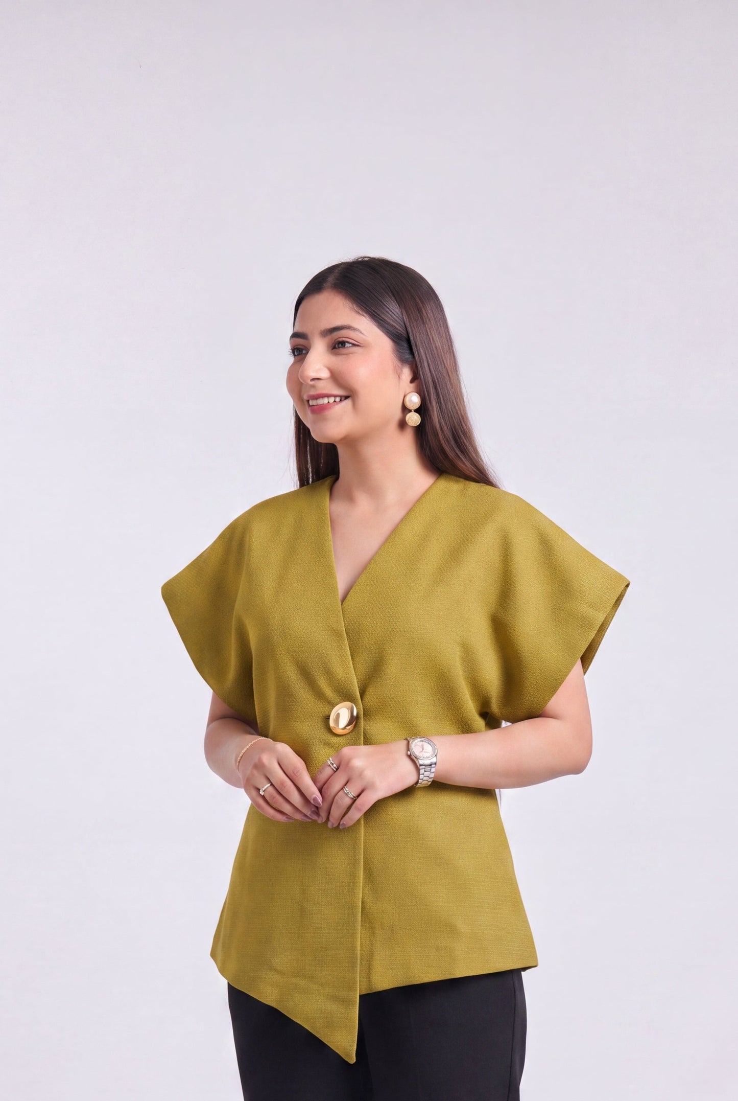 A woman modeling a mustard yellow wrap-style blouse with asymmetrical draped sleeves and black trousers, standing against a plain light gray studio background.