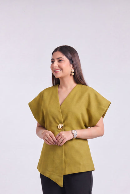 A woman modeling a mustard yellow wrap-style blouse with asymmetrical draped sleeves and black trousers, standing against a plain light gray studio background.