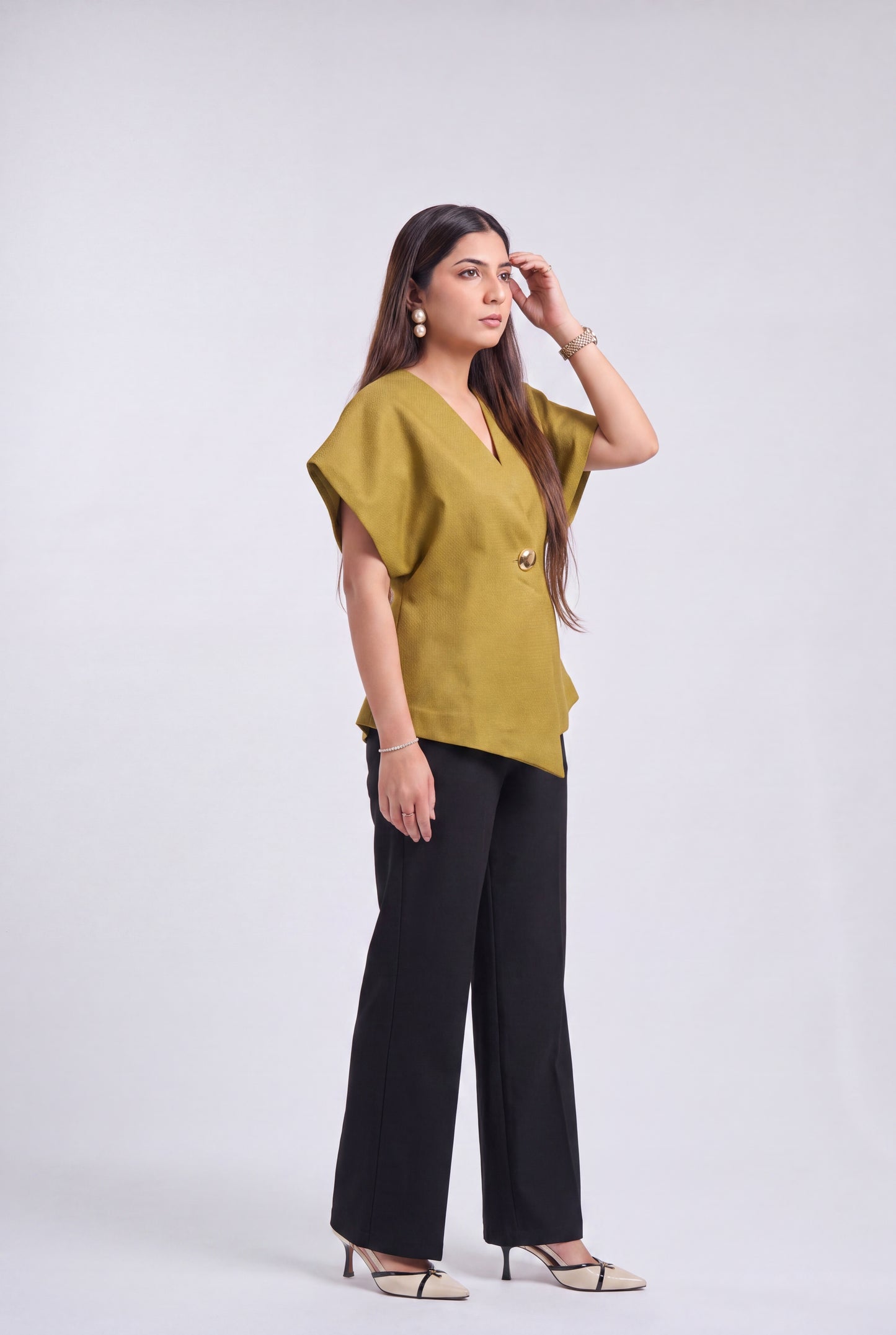 A woman modeling a mustard yellow short-sleeve top with black wide-leg pants and beige heeled sandals, standing against a light gray studio background.
