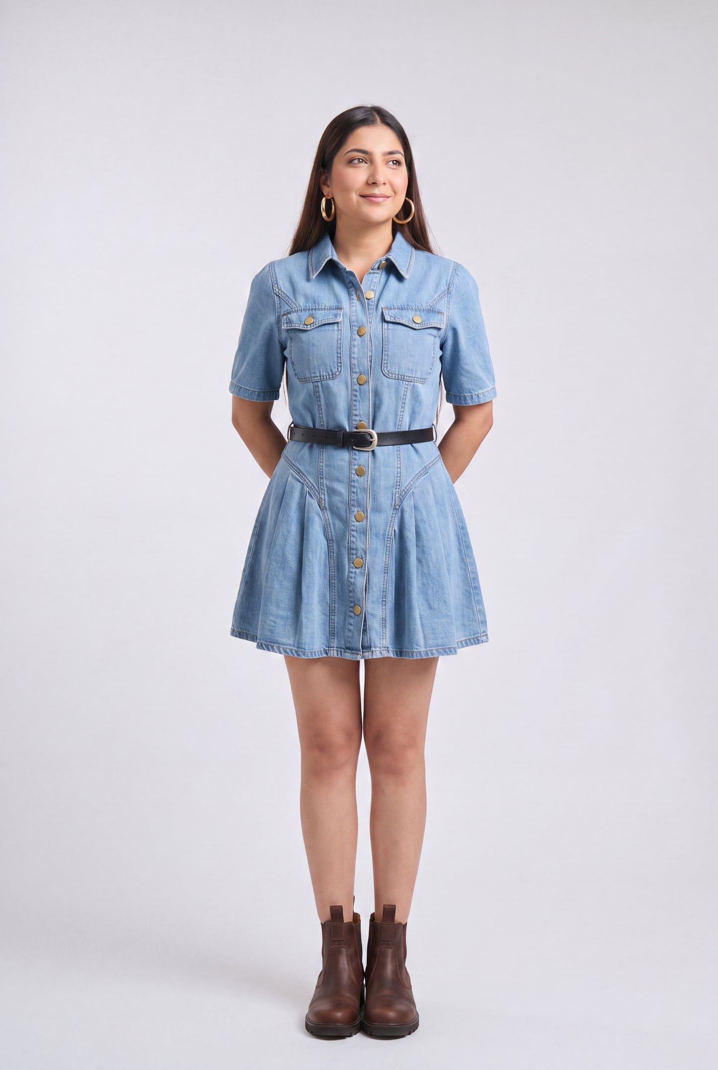 A woman modeling a light blue denim shirt dress with short sleeves, a button-down front, a black belt, and brown ankle boots, standing against a light gray studio background.