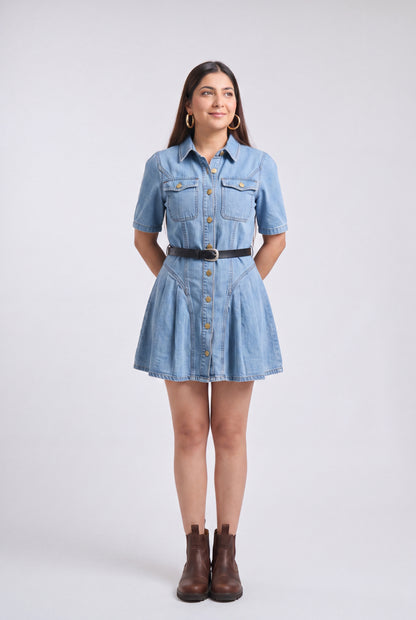 A woman modeling a light blue denim shirt dress with short sleeves, a button-down front, a black belt, and brown ankle boots, standing against a light gray studio background.