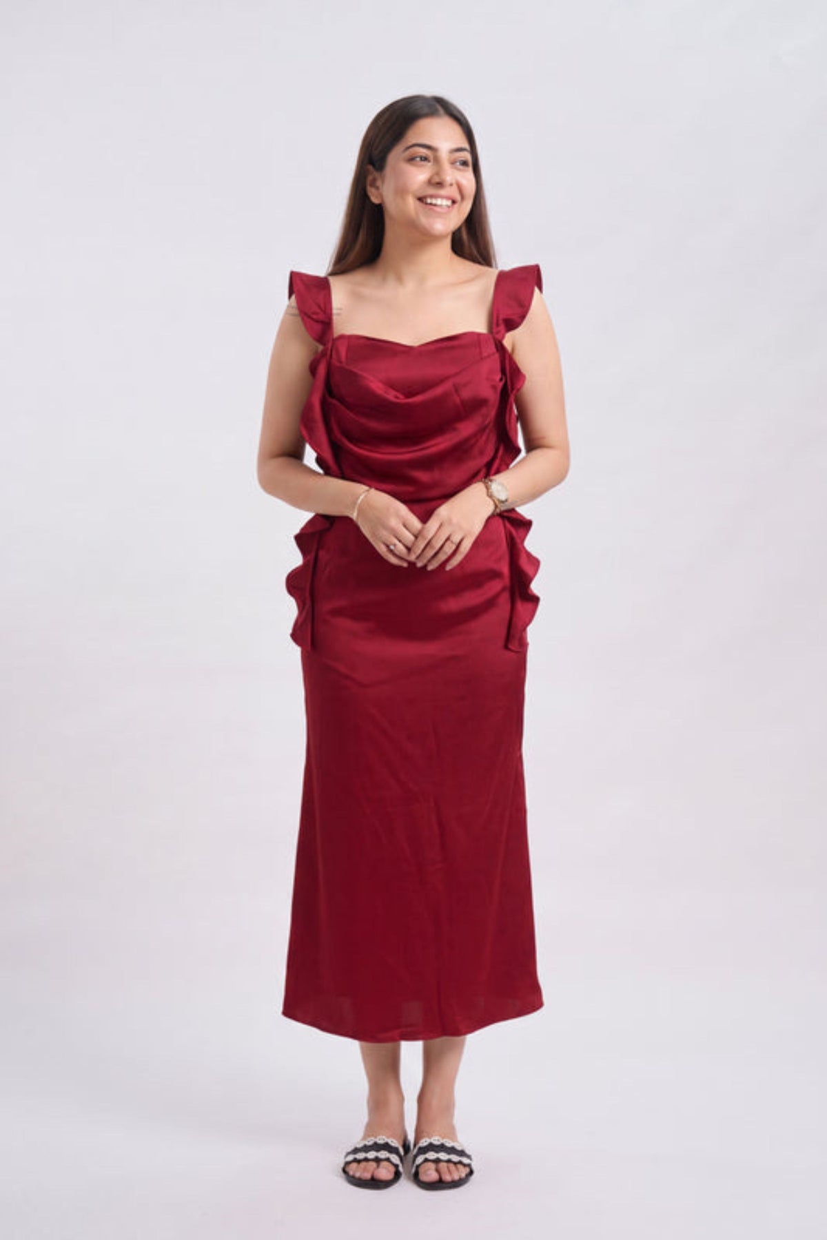 A woman modeling a sleeveless, deep red midi dress with ruffled shoulder details and a draped bodice, standing against a plain light gray background.