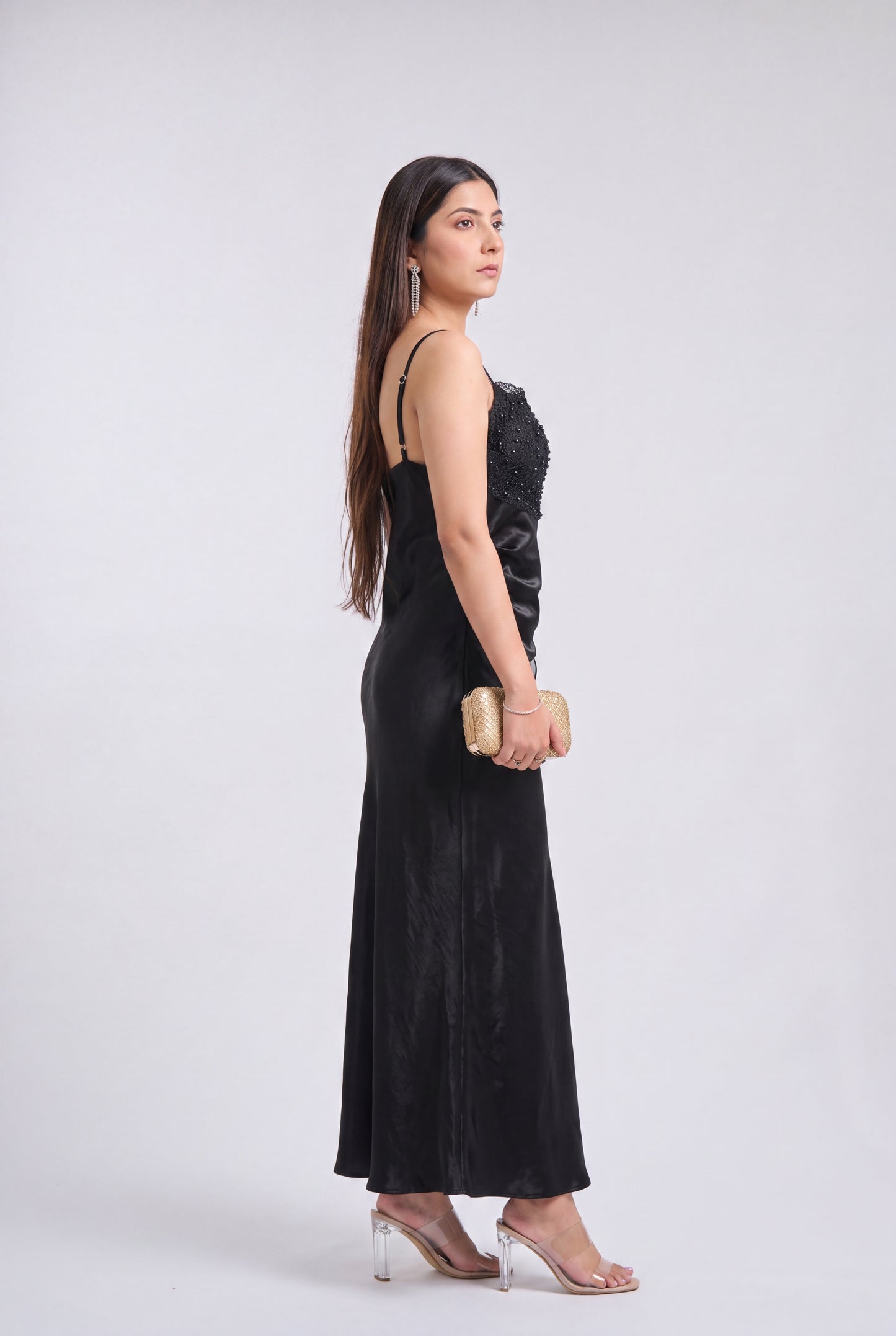 A woman stands in profile wearing a long black evening gown with a textured bodice and thin straps, holding a small beige clutch, against a light gray studio background.