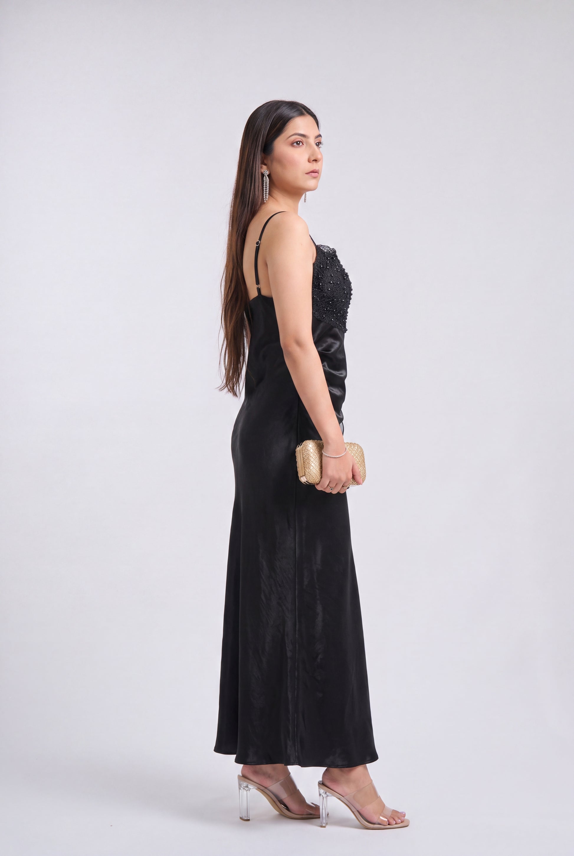 A woman stands in profile wearing a long black evening gown with a textured bodice and thin straps, holding a small beige clutch, against a light gray studio background.
