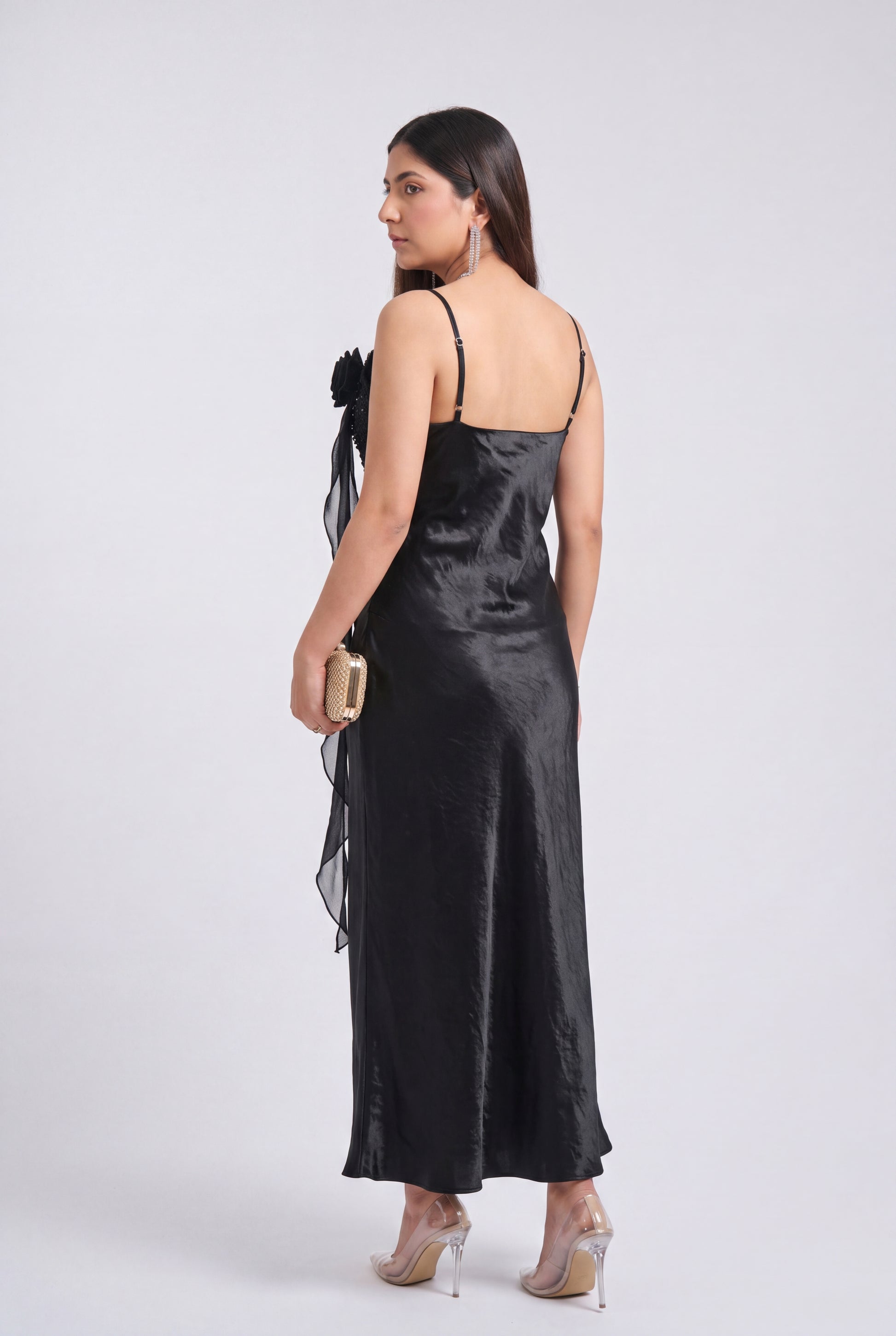 A woman modeling a black satin slip dress with thin straps, a sheer side drape, and a draped waist, shown from the back against a light gray studio background.