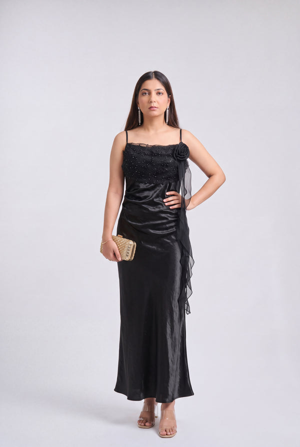 A woman modeling a black satin evening gown with spaghetti straps, a beaded bodice, and a sheer draped side detail, holding a small gold clutch against a light gray studio background.