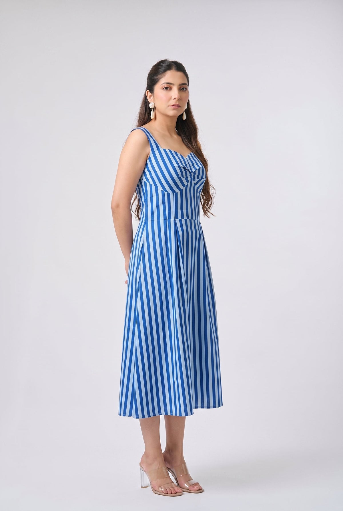 A woman modeling a sleeveless midi dress with vertical blue and white stripes, standing against a light gray studio background.