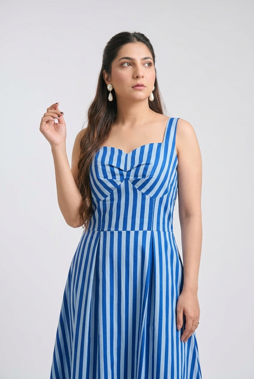 A woman modeling a sleeveless blue and white vertically striped dress with a sweetheart neckline and a bow detail at the bust, set against a plain light gray studio background.