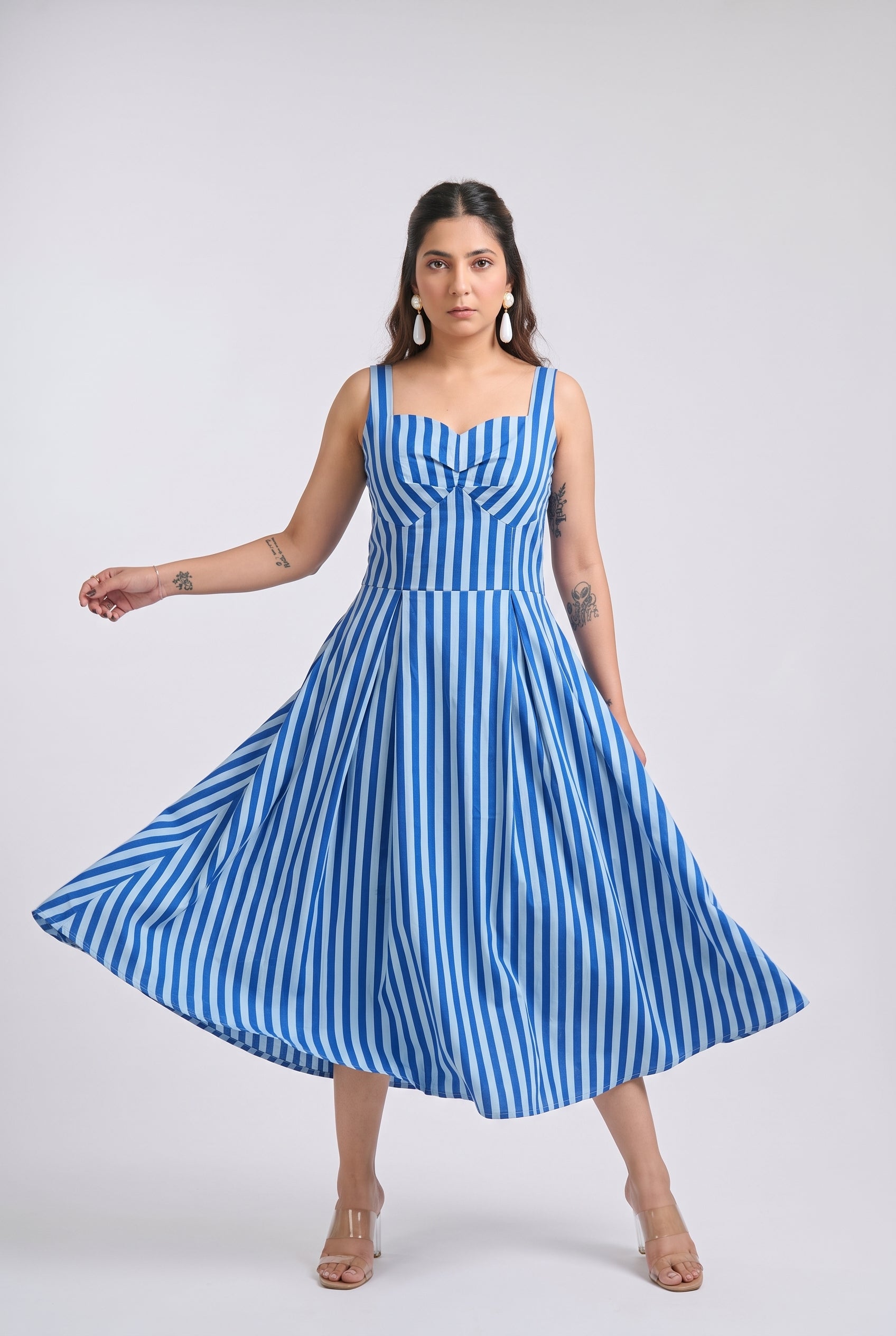A woman modeling a sleeveless blue and white vertically striped midi dress with a sweetheart neckline, gathered bodice, and flared skirt, posed against a light gray studio background.
