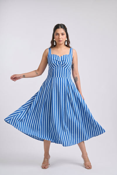 A woman modeling a sleeveless blue and white vertically striped midi dress with a sweetheart neckline, gathered bodice, and flared skirt, posed against a light gray studio background.