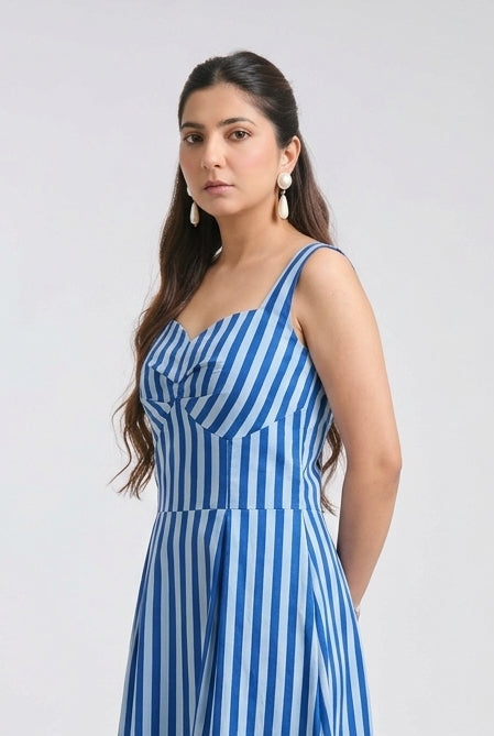 A woman modeling a sleeveless midi dress with vertical blue and white stripes, standing against a plain light gray studio background.