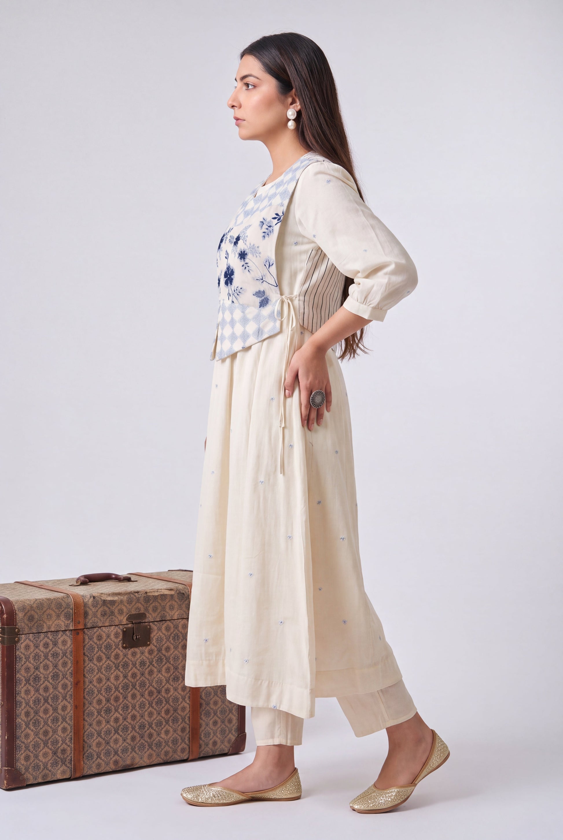 A woman models a cream-colored embroidered kurta set with a matching embroidered vest, standing next to a vintage patterned trunk against a light gray studio background.