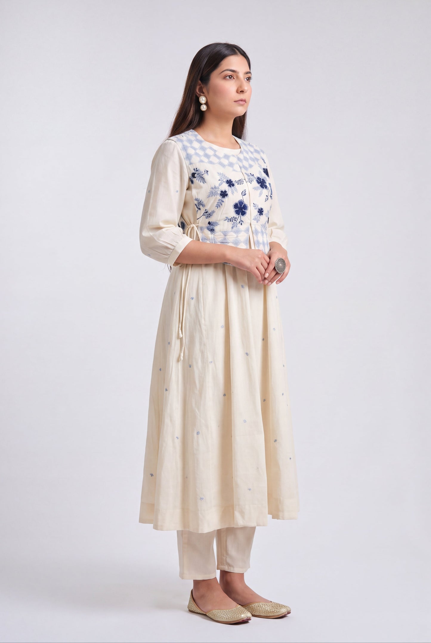A woman modeling a cream-colored kurta set with a blue and white checkered yoke, blue floral embroidery, and small blue embroidered details on the skirt, paired with matching cream pants and beige flats, against a light gray studio background.