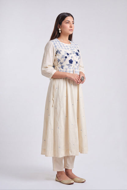 A woman modeling a cream-colored kurta set with a blue and white checkered yoke, blue floral embroidery, and small blue embroidered details on the skirt, paired with matching cream pants and beige flats, against a light gray studio background.
