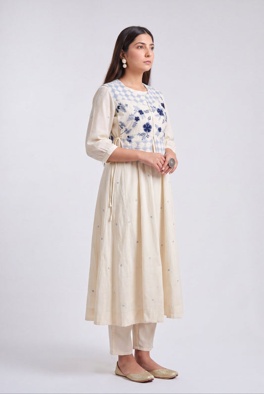 A woman modeling a cream-colored kurta set with a blue and white checkered yoke, blue floral embroidery, and small blue embroidered details on the skirt, paired with matching cream pants and beige flats, against a light gray studio background.