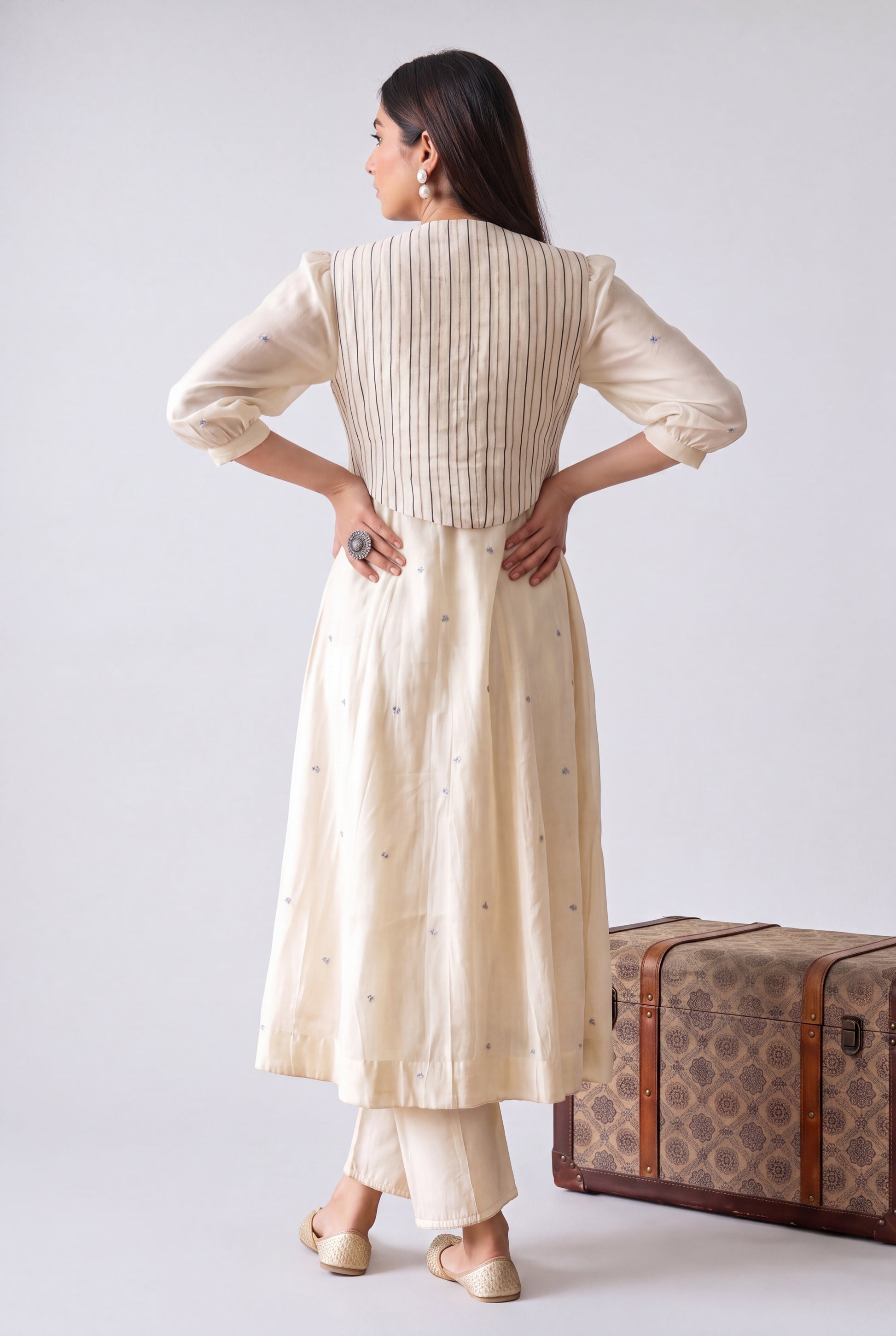 A woman stands with her back to the camera wearing a cream-colored kurta with vertical pleats and subtle embroidered details, paired with matching pants. She is posed next to a vintage-style patterned trunk against a plain light gray studio background.