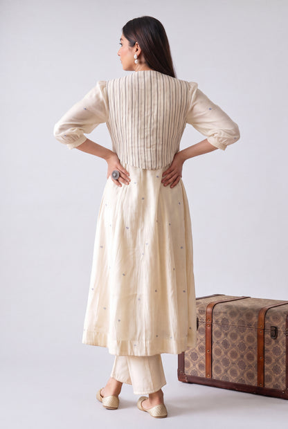 A woman stands with her back to the camera wearing a cream-colored kurta with vertical pleats and subtle embroidered details, paired with matching pants. She is posed next to a vintage-style patterned trunk against a plain light gray studio background.