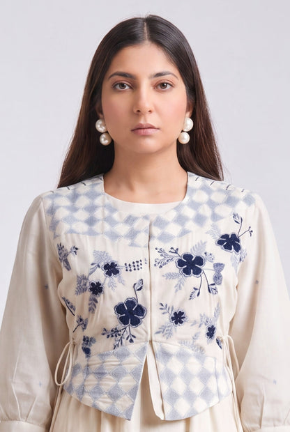 A woman stands against a light gray studio background wearing a cream-colored traditional outfit with a blue and white gingham embroidered vest, matching pants, and beige flat shoes.