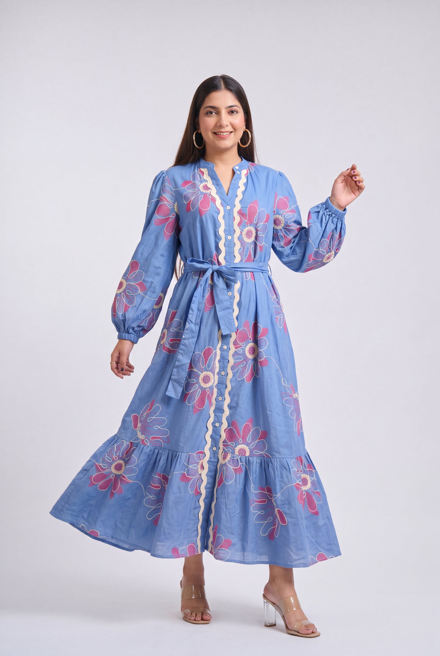 A woman modeling a light blue maxi dress with pink and white floral embroidery, a cream lace trim, and a matching waist tie, set against a plain light gray studio background.