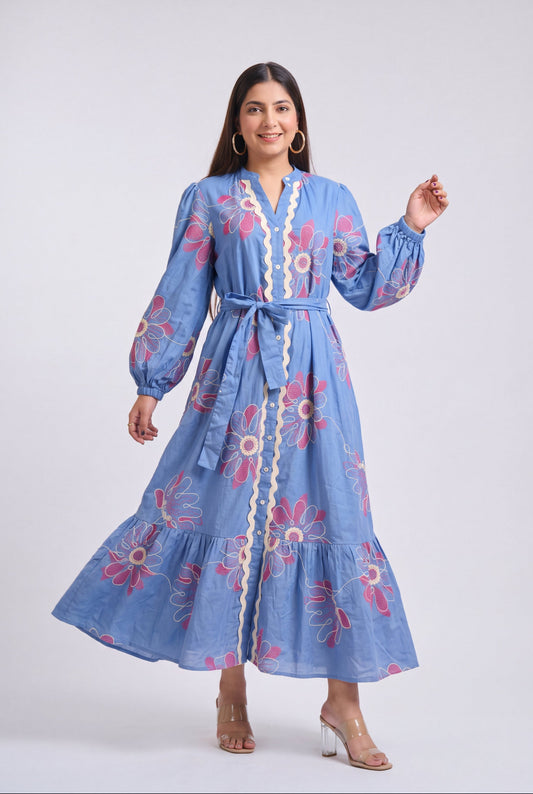 A woman modeling a light blue maxi dress with pink and white floral embroidery, a cream lace trim, and a matching waist tie, set against a plain light gray studio background.